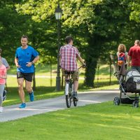 Family jogging and man cycling in park, people walking in background.